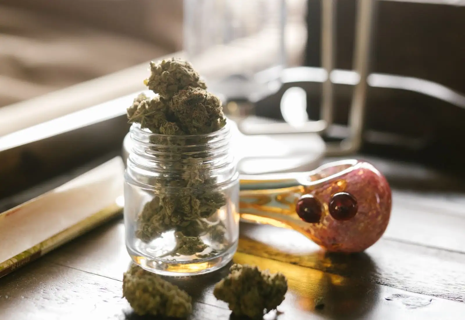 Bocal en verre rempli de fleurs de chanvre séchées, posé à côté d'une pipe en verre et d'un carnet de feuilles à rouler