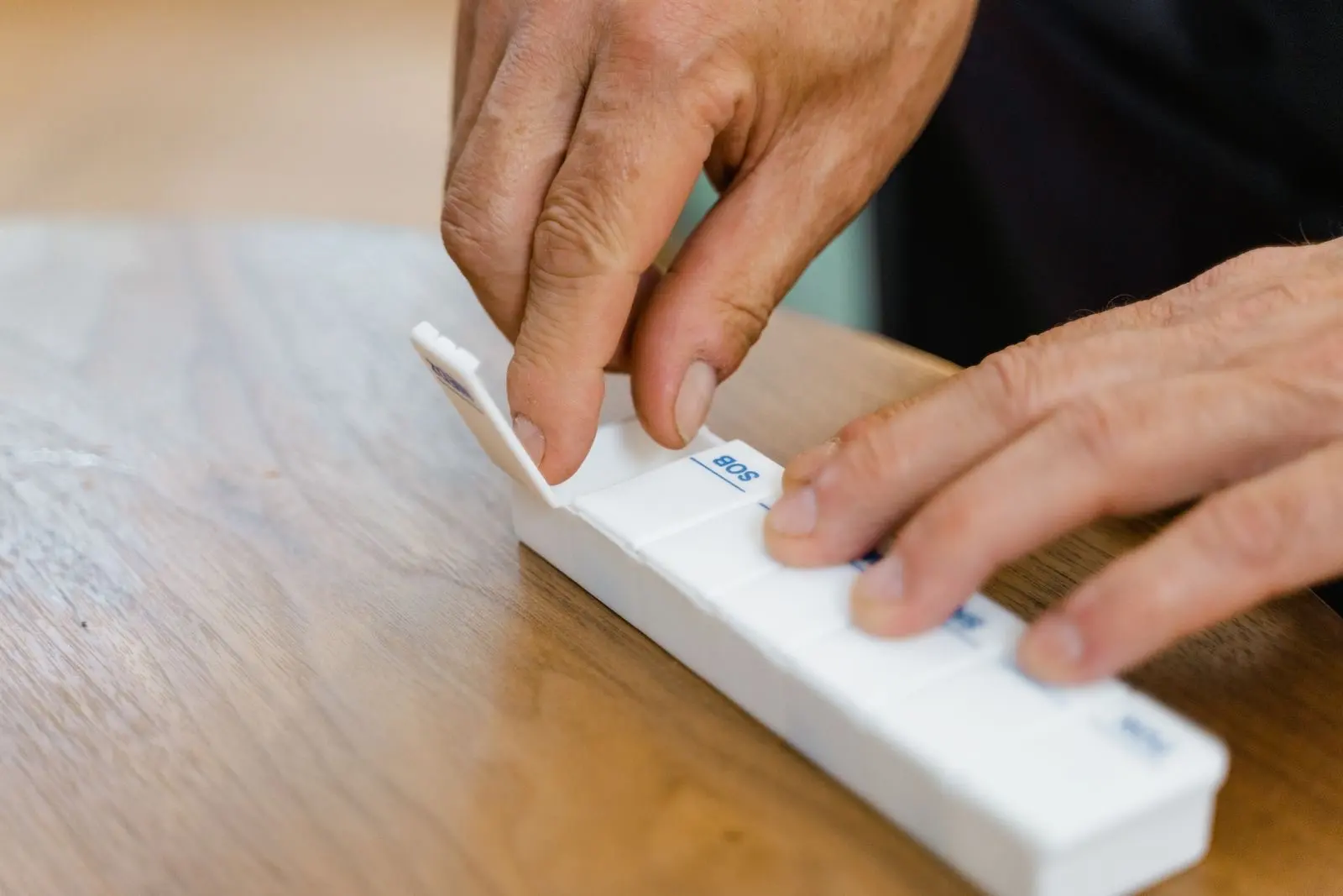 Mains d'une personne âgée qui prépare ses médicaments dans un pilulier hebdomadaire posé sur une table en bois