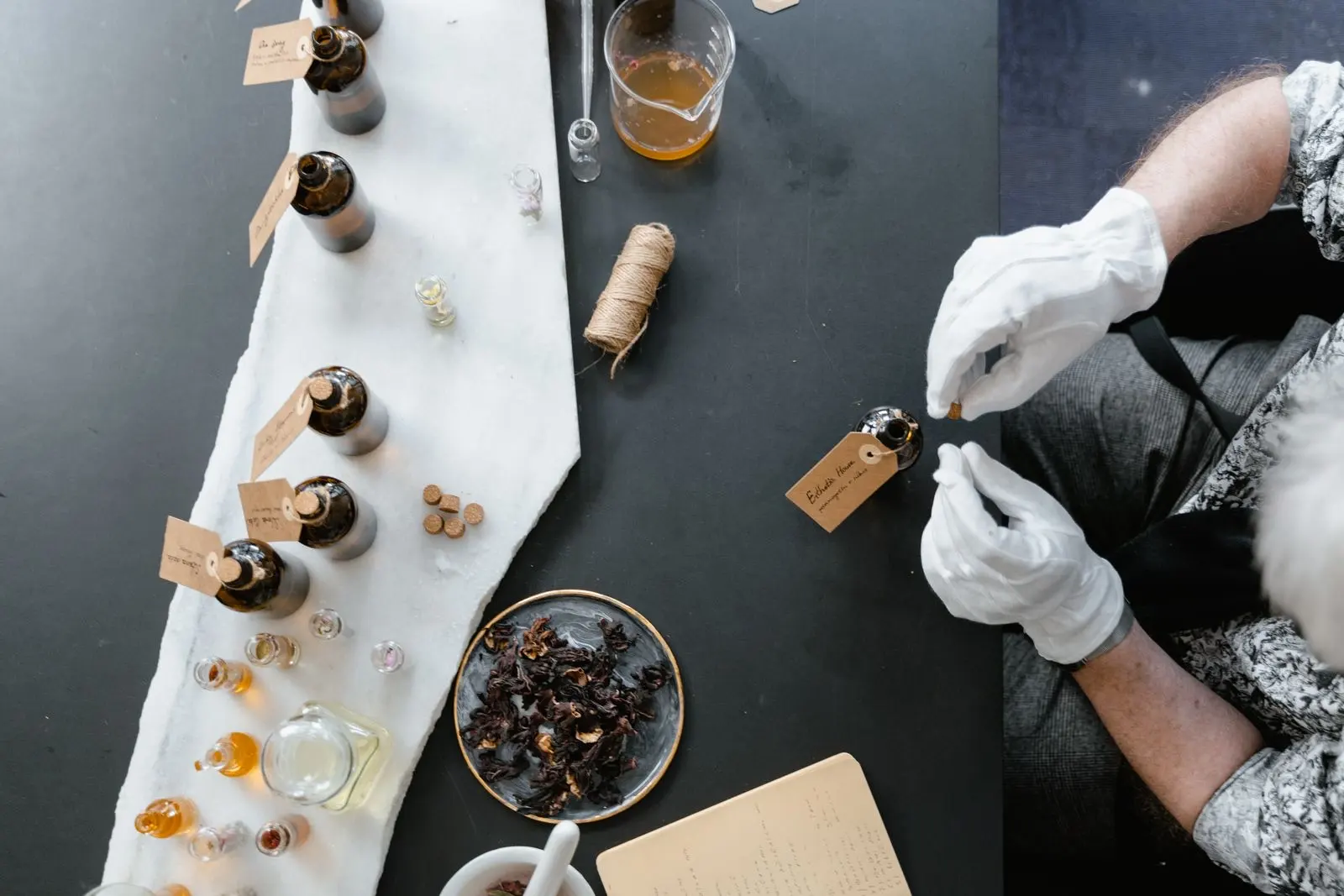 Vue de dessus d'un atelier de parfumeur : flacons de matières premières étiquetés, fleurs séchées et mains gantées qui préparent une composition