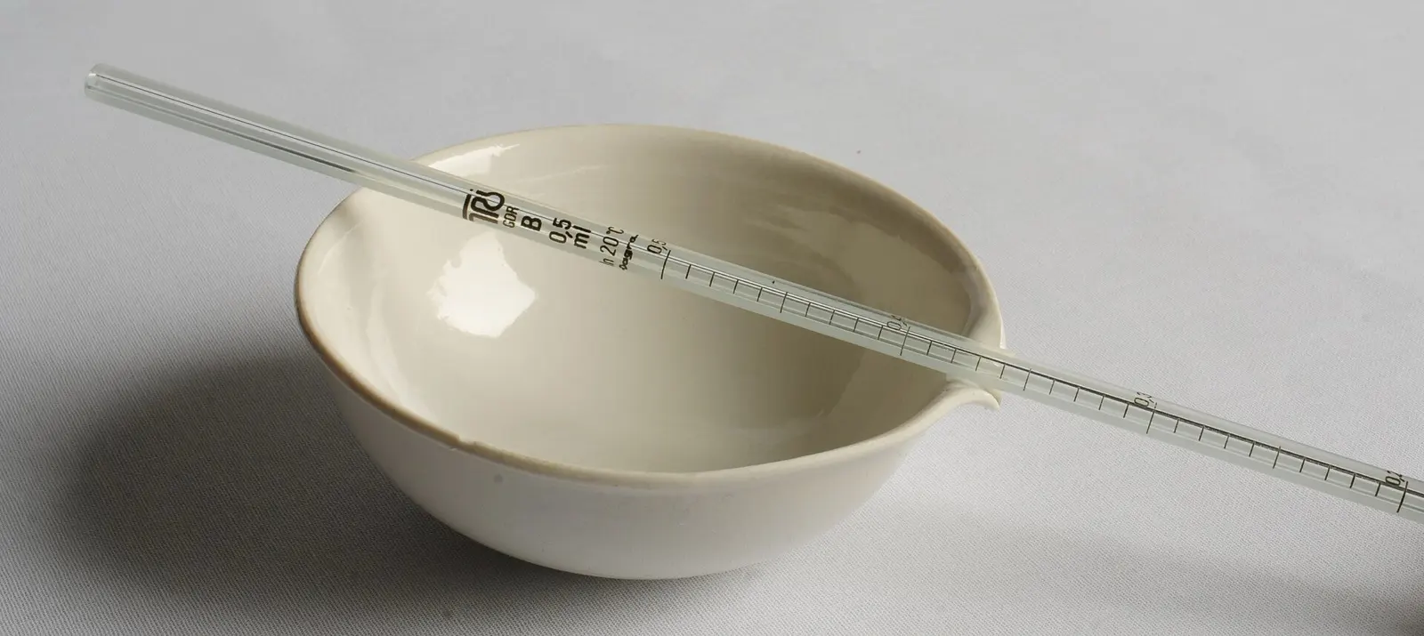 Pipette graduée en verre de 0,5 ml posée en travers d'un petit bol blanc en porcelaine, sur un fond gris clair