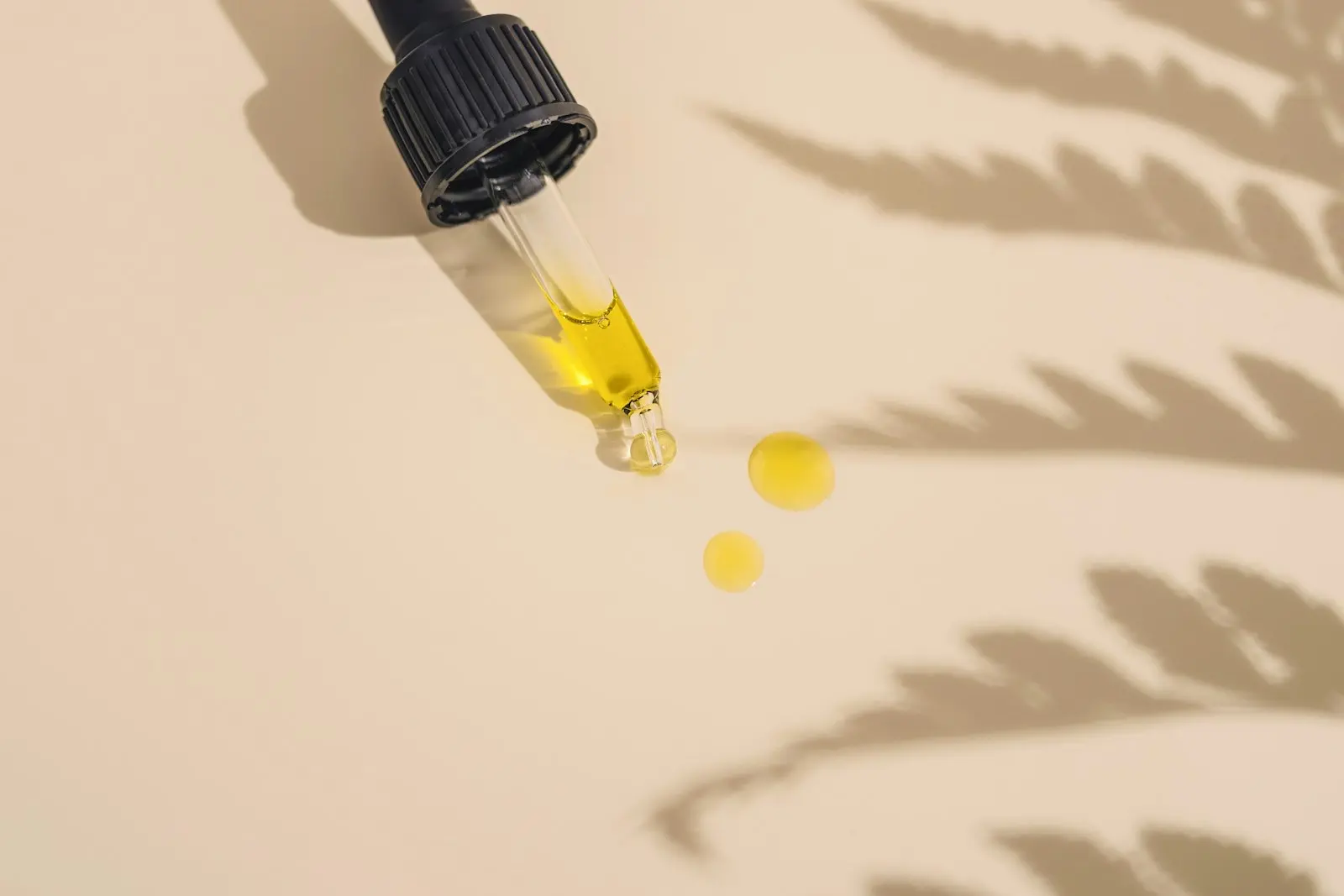 Pipette de flacon compte-gouttes remplie d'une huile dorée, deux gouttes posées sur un fond beige