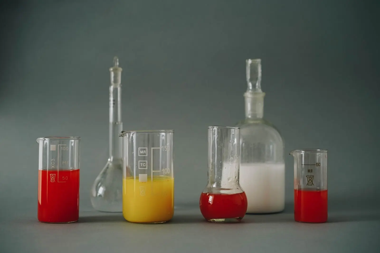 Béchers et fioles de laboratoire remplis de liquides rouges, jaunes et blancs sur fond gris