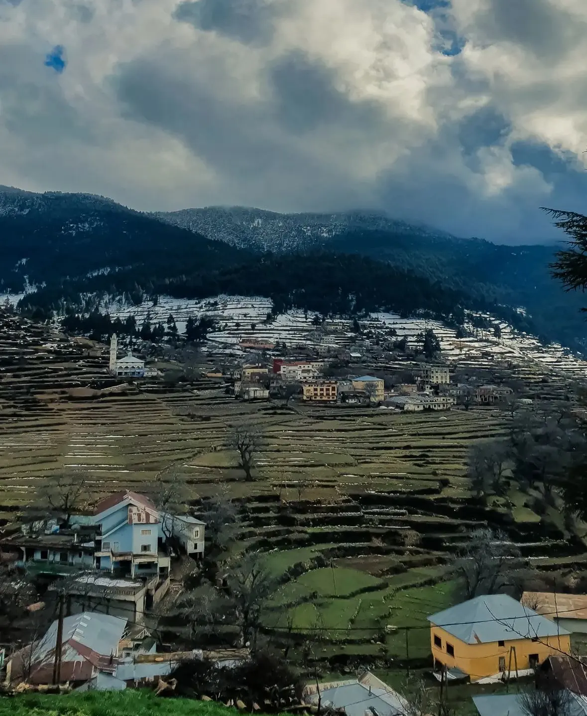 Champs en terrasses autour du village de Kétama, dans le Rif, avec un minaret, des maisons colorées et des plaques de neige sous un ciel chargé
